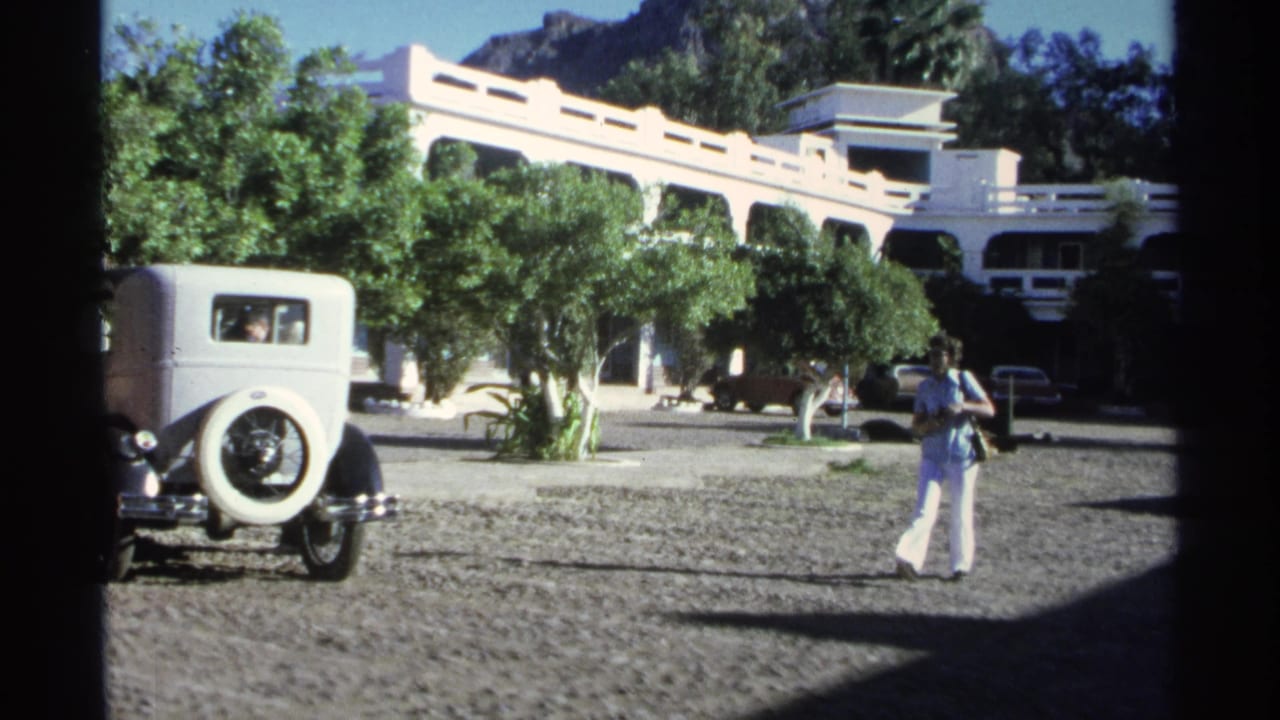 1975 Guaymas home-movie frame with a woman in a blue top and white pants standing near parked cars outside a white arched building.