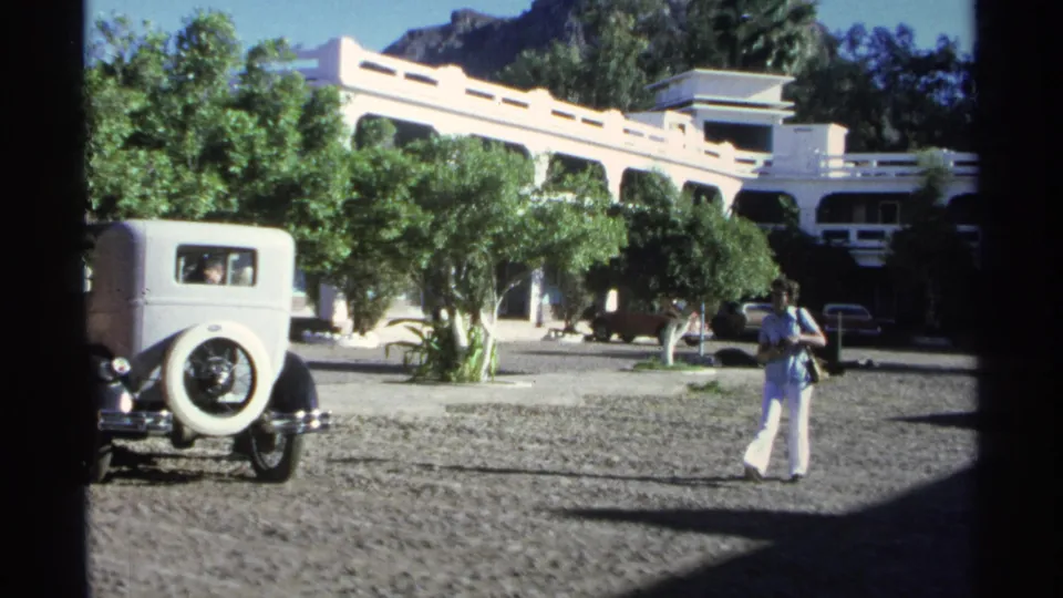 1975 Guaymas home-movie frame with a woman in a blue top and white pants standing near parked cars outside a white arched building.