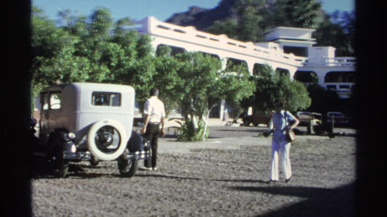 1975 Guaymas frame showing two adults near a white vintage sedan, with trees, gravel, parked cars, and a white arched building behind them.