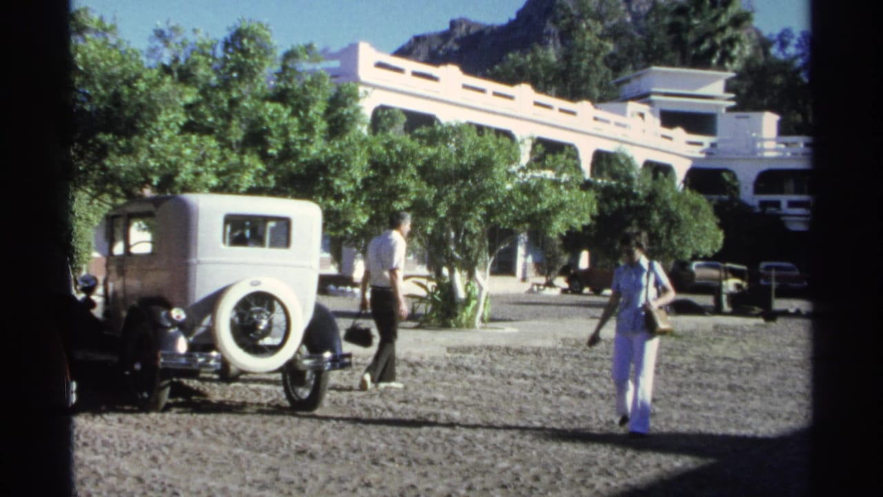 In Guaymas, Mexico, 1975, two adults walk through a gravel lot beside a white vintage sedan, trees, and a white building.
