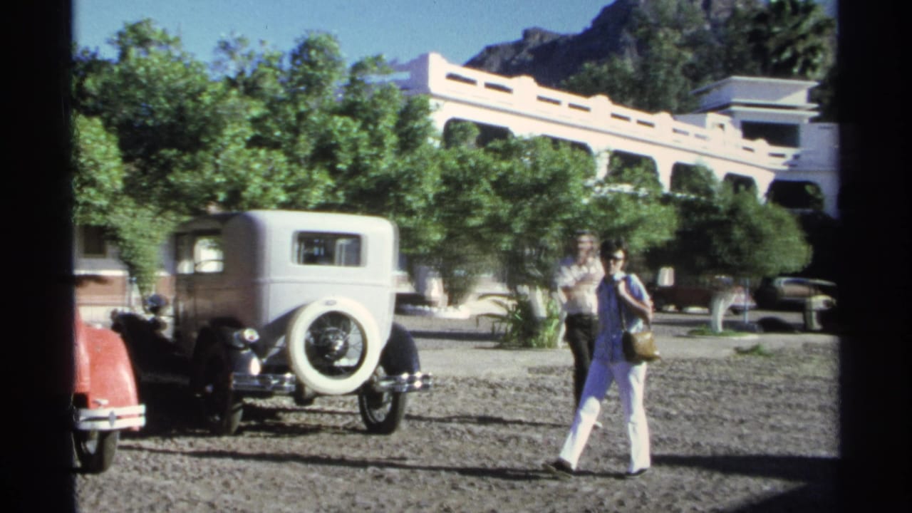 Woman walks past a white vintage sedan as a man stands or walks nearby in the lot.