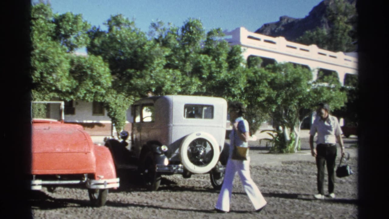 In the 1975 Guaymas home-movie frame, the adults pass each other near a white sedan, a red car, trees, and a white building.