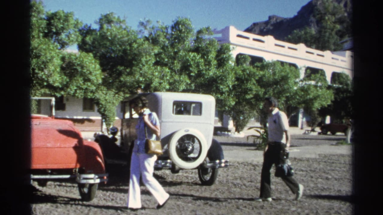 The woman continues past the white sedan while the man moves the other direction across the Guaymas gravel parking area.