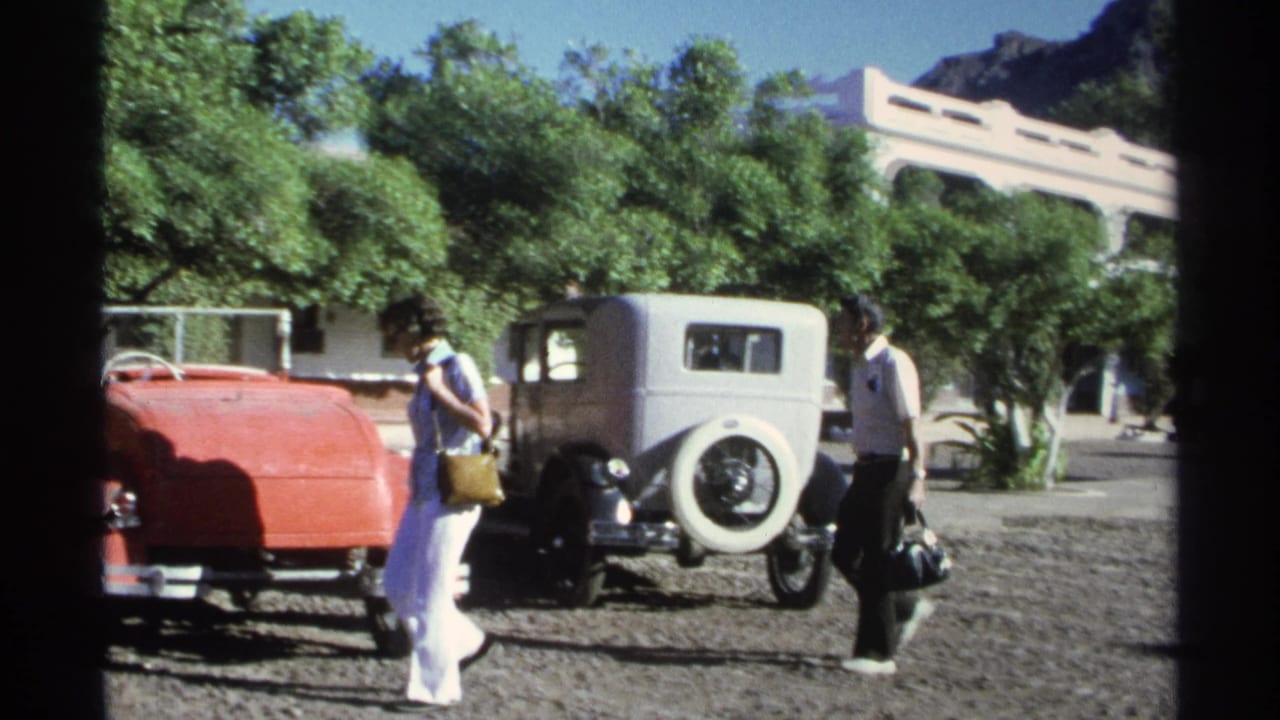 Two adults walk across a gravel lot toward parked vintage cars under trees.