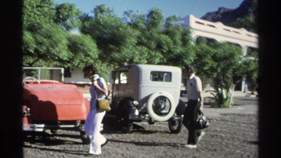In Guaymas, Mexico in 1975, two adults approach a red convertible and light gray sedan in a sunny gravel lot.