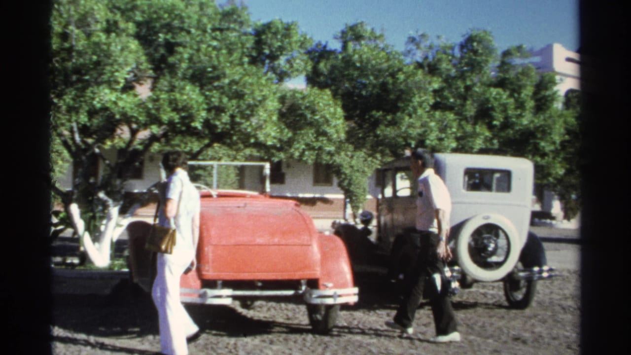 Two adults in 1975 Guaymas walk near a red vintage convertible and gray sedan, with trees and a building behind them.