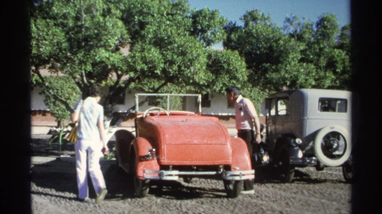 The adults pause near the rear of a red convertible in Guaymas, Mexico, with a gray sedan, trees, and gravel visible.