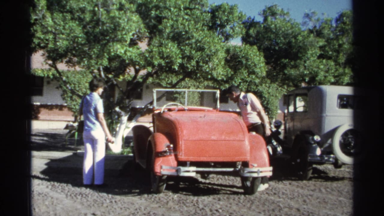 Adults stand near a red convertible and gray sedan on a gravel lot under trees.
