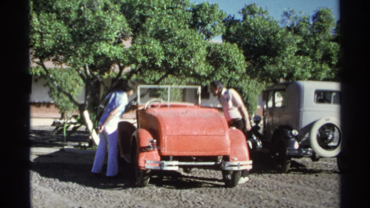 Adults inspect a red convertible in a Guaymas gravel lot, with a gray vintage sedan parked close behind.