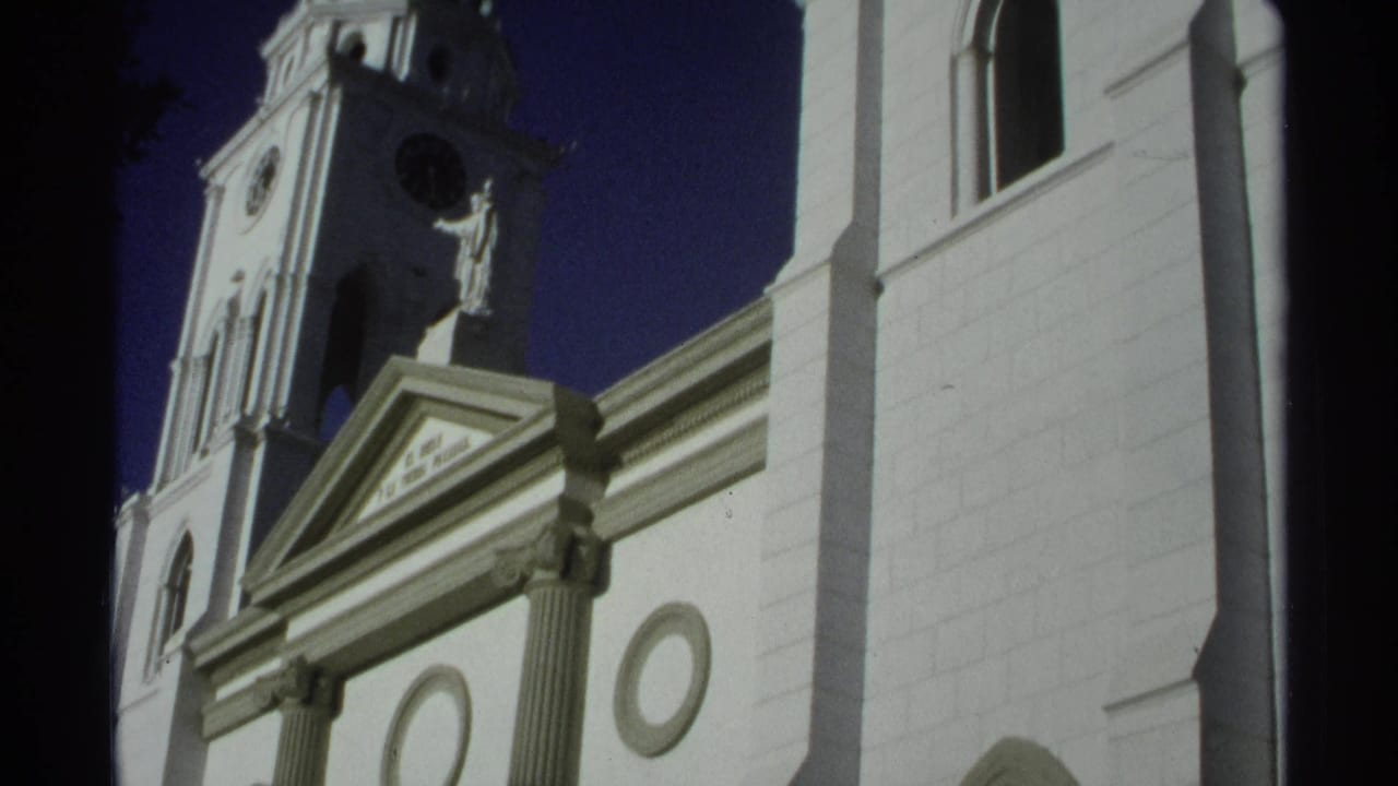 Low-angle white church facade with clock tower, statue, columns, and deep blue sky.