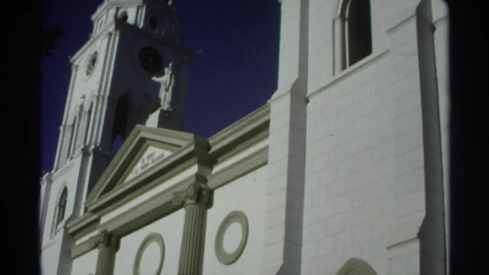 1975 Guaymas, Mexico 8mm frame showing a low-angle white church facade with clock tower, statue, pediment, and blue sky.