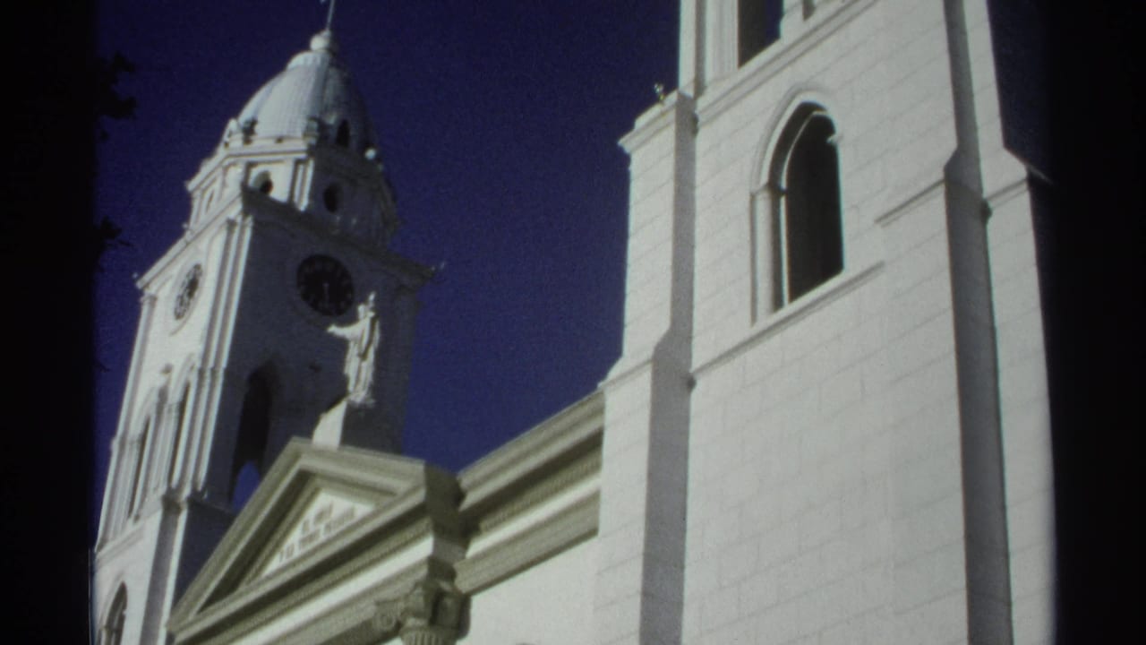 1975 Guaymas, Mexico frame of a white church exterior, showing a clock tower, cross-topped dome, and tall arched facade opening.