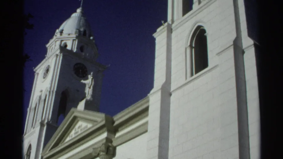1975 Guaymas, Mexico frame of a white church exterior, showing a clock tower, cross-topped dome, and tall arched facade opening.