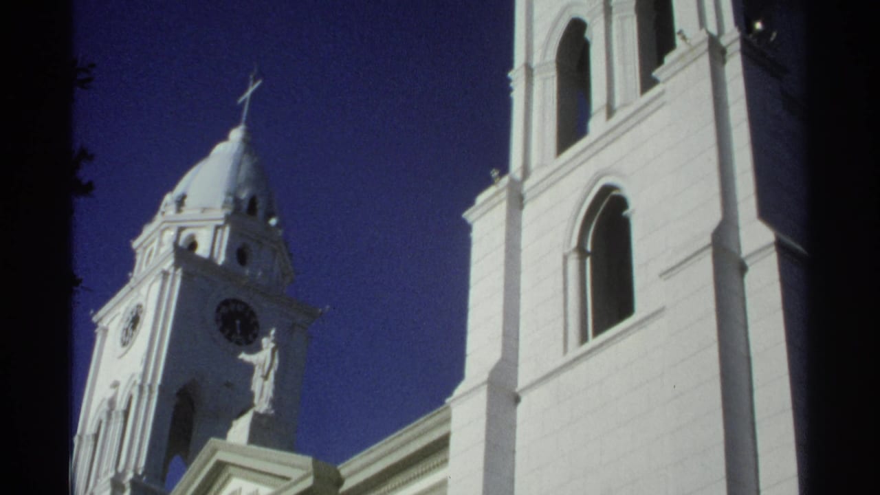 1975 Guaymas, Mexico 8mm frame with an upward view of the white church clock tower, cross, statue, and adjacent arched facade.
