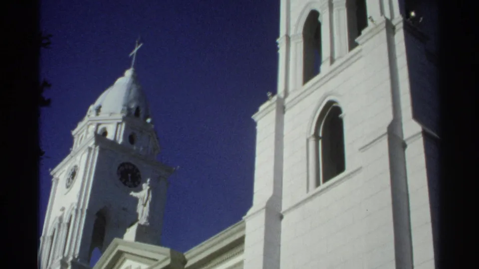 1975 Guaymas, Mexico 8mm frame with an upward view of the white church clock tower, cross, statue, and adjacent arched facade.