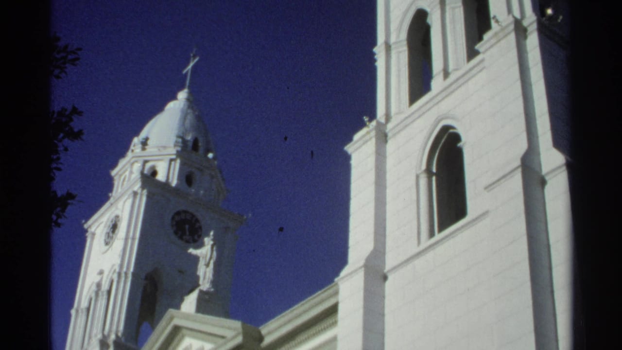 1975 Guaymas, Mexico frame showing the church tower with clock, facade statue, cross, and adjoining white wall with arched opening.