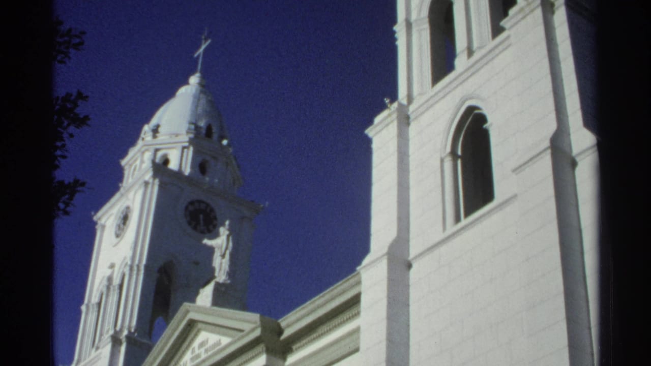 Low-angle view of a white church tower with clock, dome, cross, and statue against a dark blue sky.