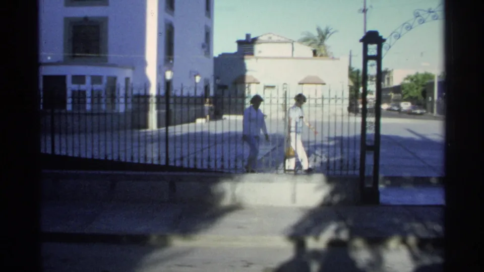 1975 Guaymas street-level church view through a gate, with two women in light clothing walking across the entrance area.