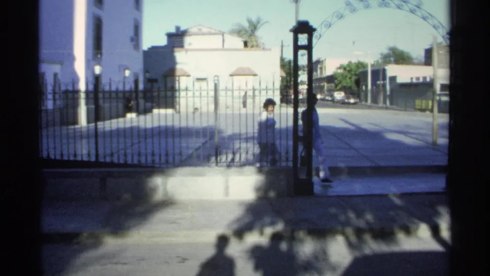 1975 Guaymas church entrance seen through wrought iron, with women near the gate and sunlit buildings across the street.