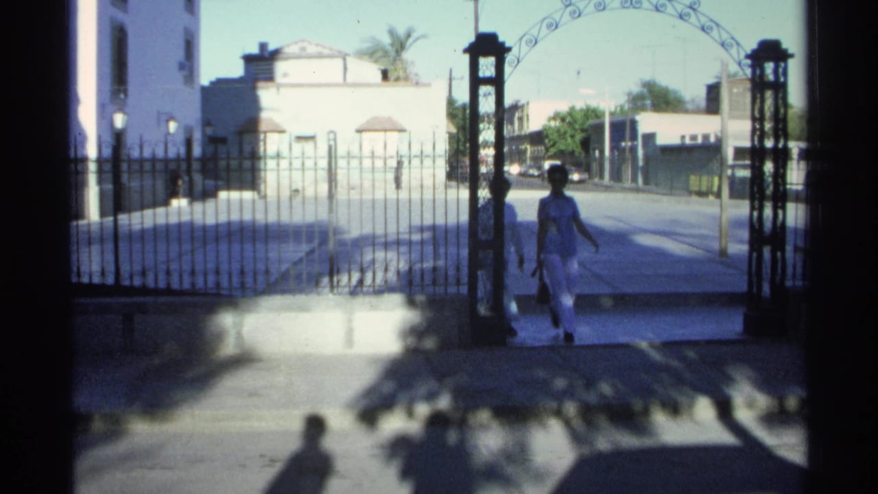 In 1975 Guaymas, Mexico, two women in light clothing walk near an arched gate beside a white building and iron fence.