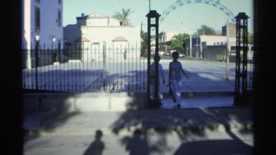 In 1975 Guaymas, Mexico, two women in light clothing walk near an arched gate beside a white building and iron fence.