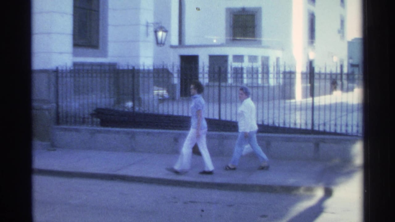A soft 8mm frame shows two women walking together along a Guaymas sidewalk beside iron fencing and a white building.