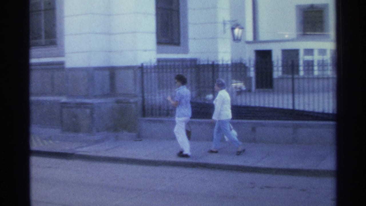 The women continue along the sunlit sidewalk in Guaymas, with the white building, iron fence, and street curb behind them.