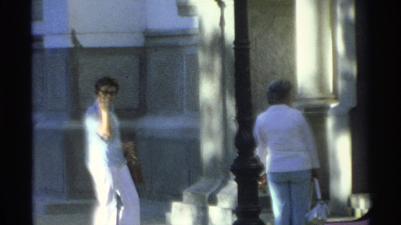 In 1975 Guaymas home-movie footage, a woman in white pants and sunglasses faces the camera near a sunlit building as another woman walks away.