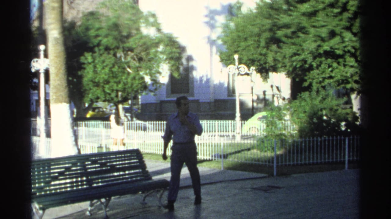A man walks through a park-like public area in Guaymas, Mexico, with a green bench, palm trees, white fencing, and strong sunlit contrast.