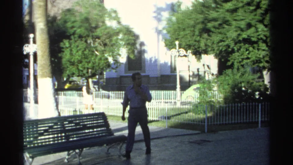 A man walks through a park-like public area in Guaymas, Mexico, with a green bench, palm trees, white fencing, and strong sunlit contrast.