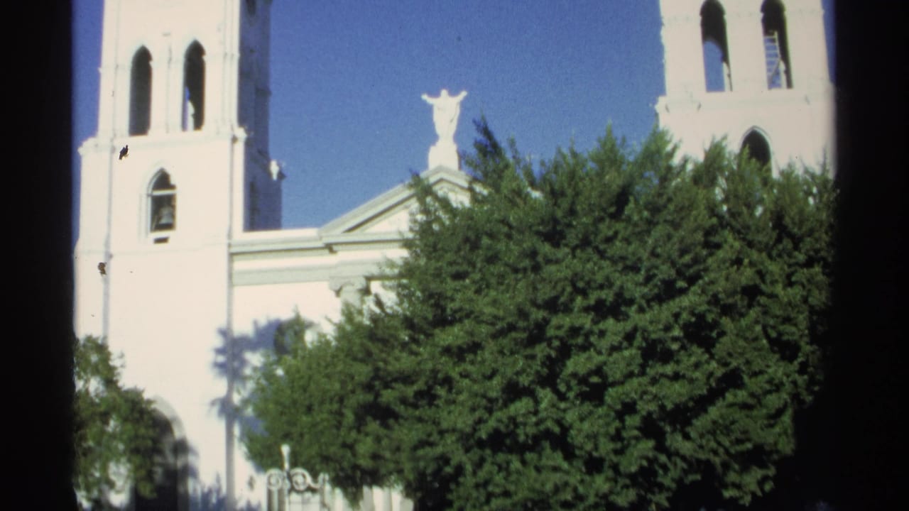 1975 8mm view in Guaymas, Mexico, showing a white church facade, bell towers, roof statue, trees, and clear blue sky.