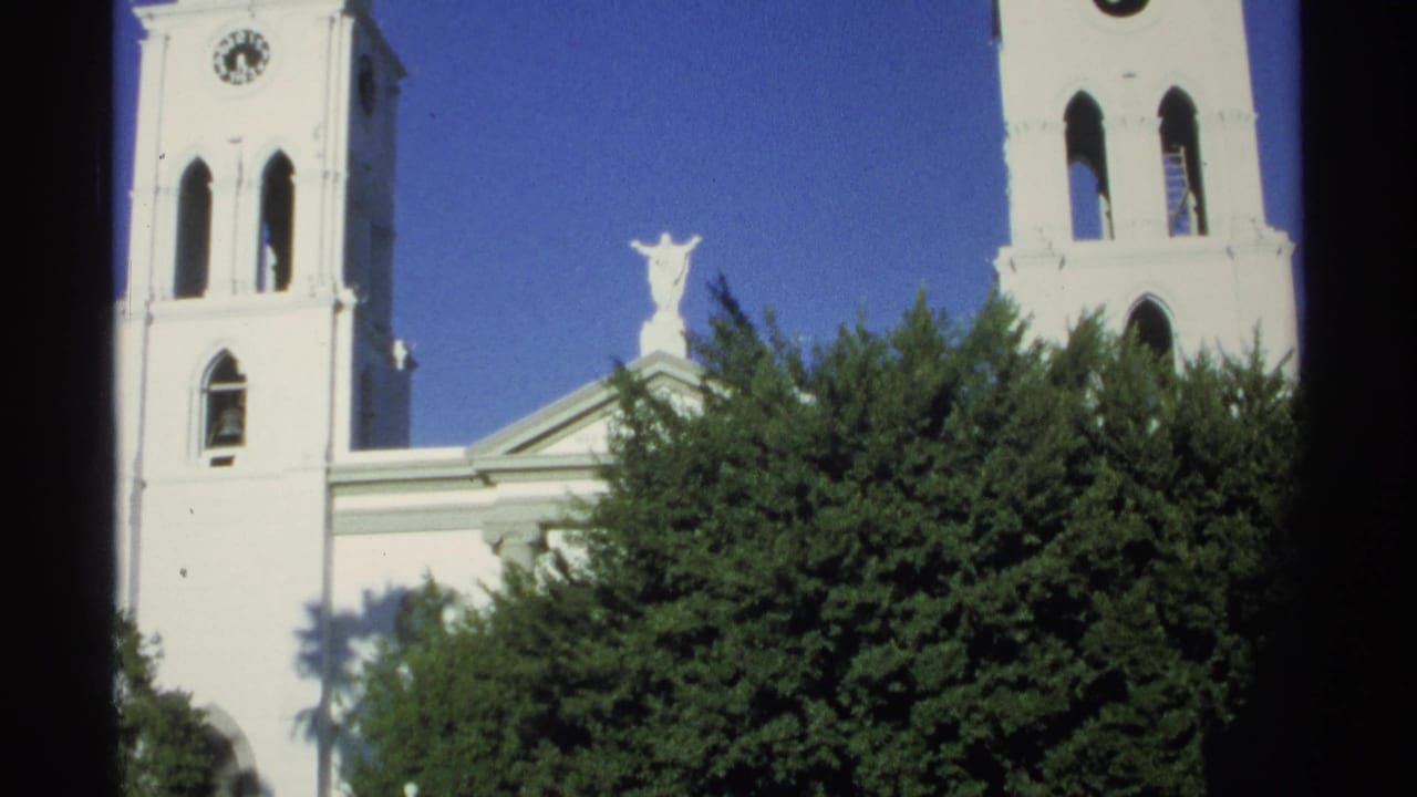 Guaymas, Mexico, 1975 home-movie frame of a white church tower, clock face, roof statue, tree canopy, and saturated blue sky.