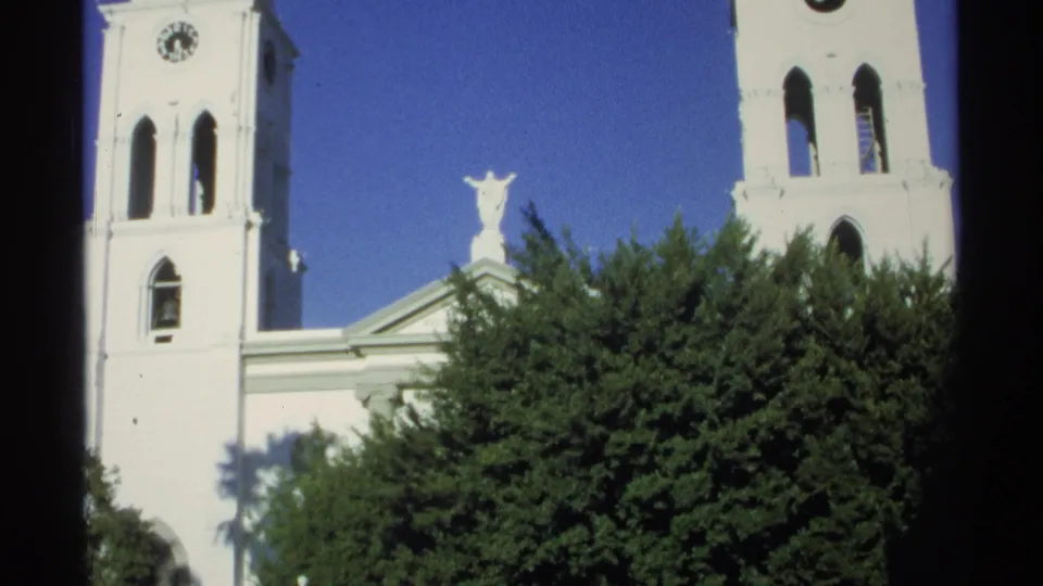 Guaymas, Mexico, 1975 home-movie frame of a white church tower, clock face, roof statue, tree canopy, and saturated blue sky.