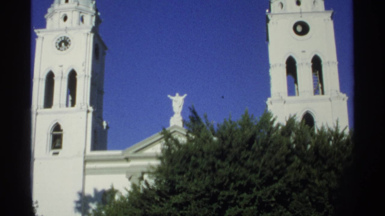 1975 Guaymas church exterior with both bell towers, a central roof statue, arched windows, green trees, and 8mm film softness.