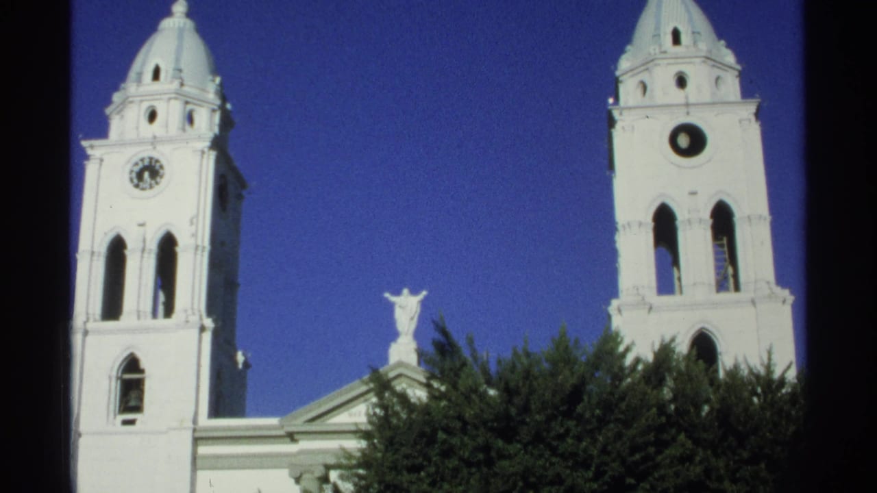 Final 1975 8mm frame of the Guaymas church exterior, with twin towers, clock face, roof statue, trees, and vivid blue sky.