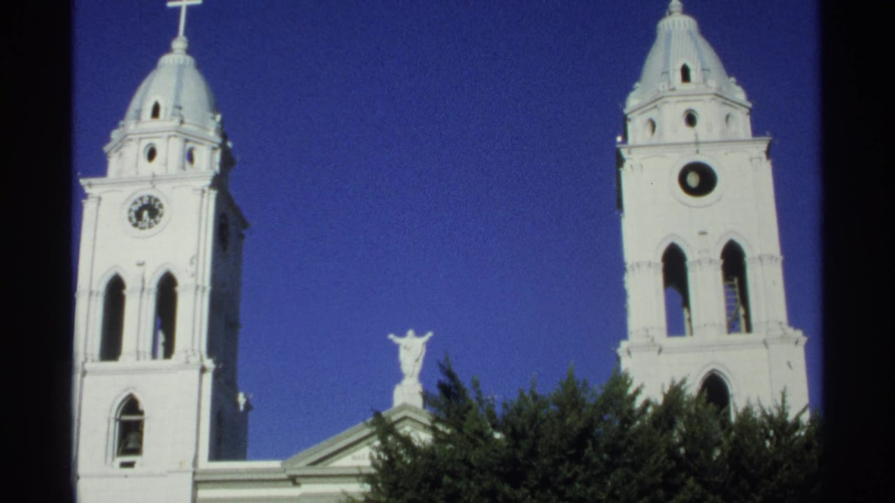 1975 Guaymas, Mexico 8mm frame showing twin white cathedral towers, clock faces, crosses, a central statue, trees, and deep blue sky.
