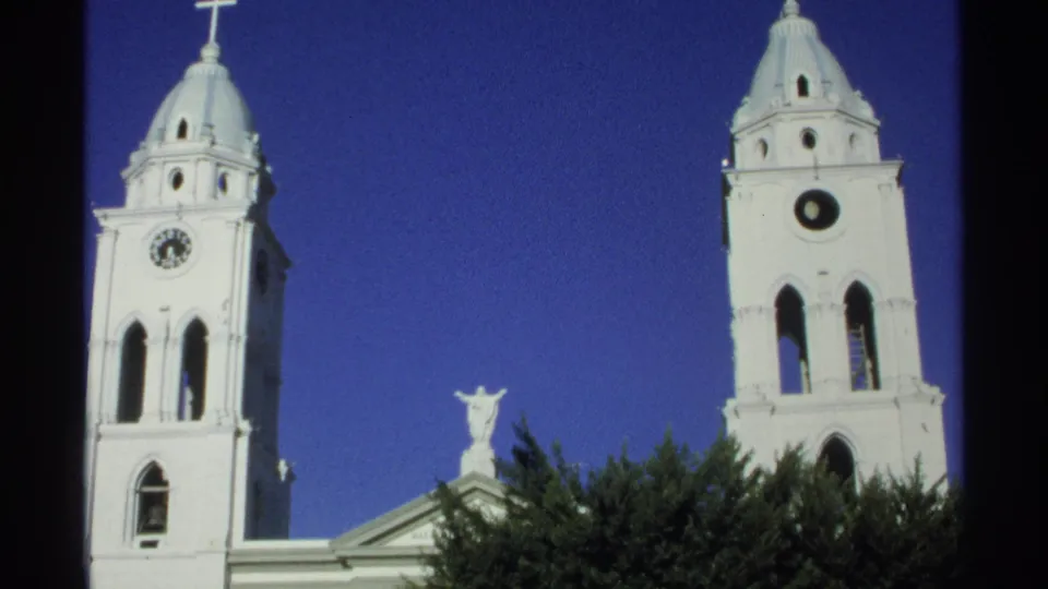 1975 Guaymas, Mexico 8mm frame showing twin white cathedral towers, clock faces, crosses, a central statue, trees, and deep blue sky.