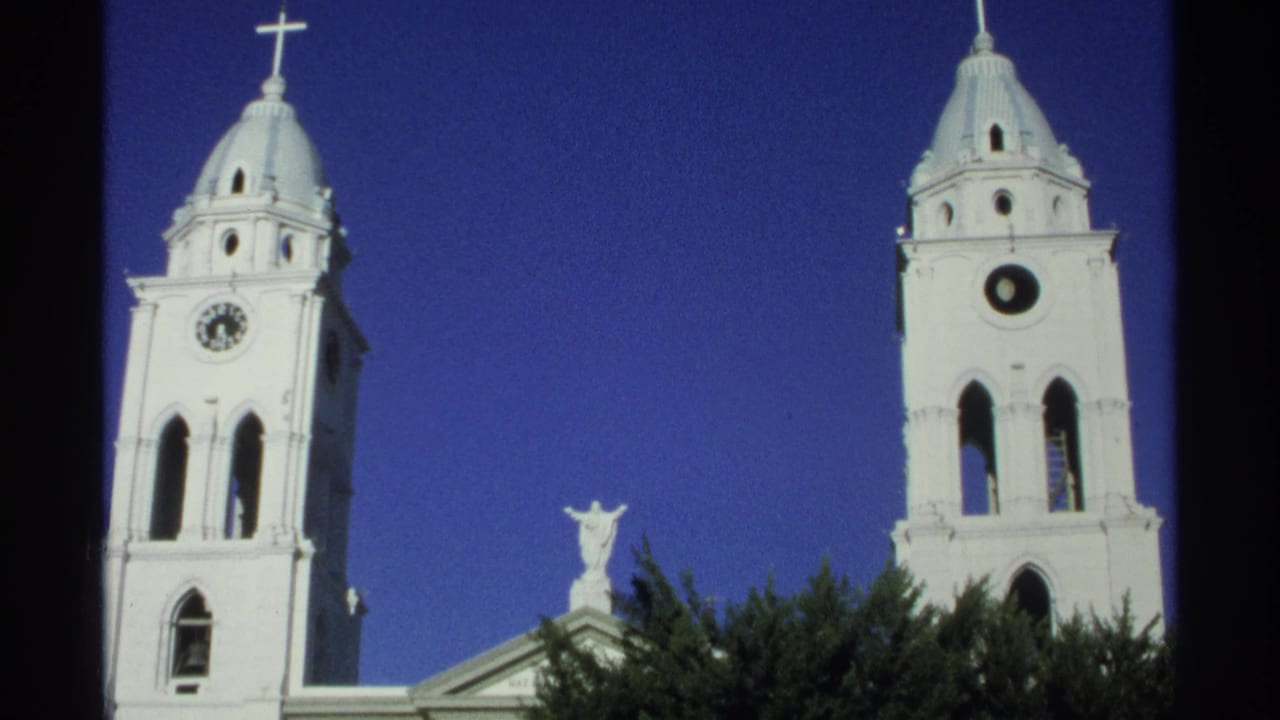 1975 Guaymas, Mexico 8mm still showing twin cathedral towers, visible clock faces, crosses, a statue between them, and trees below.