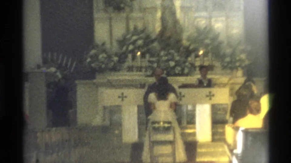 In 1975 Guaymas, Mexico, a bride and groom face a priest at a flower-decorated church altar filmed from the pews on 8mm.
