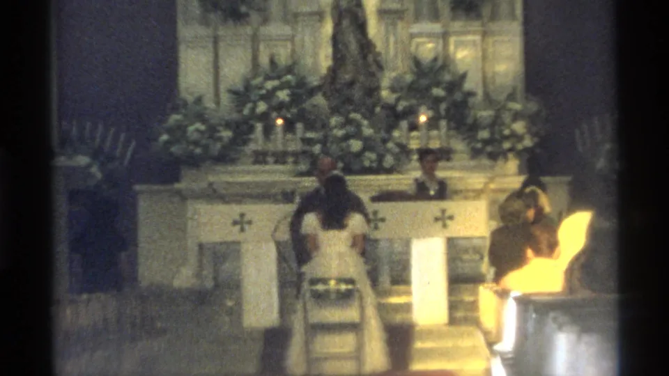 A handheld 8mm view from the pews shows the couple at the altar with candles, flowers and the priest in the background.