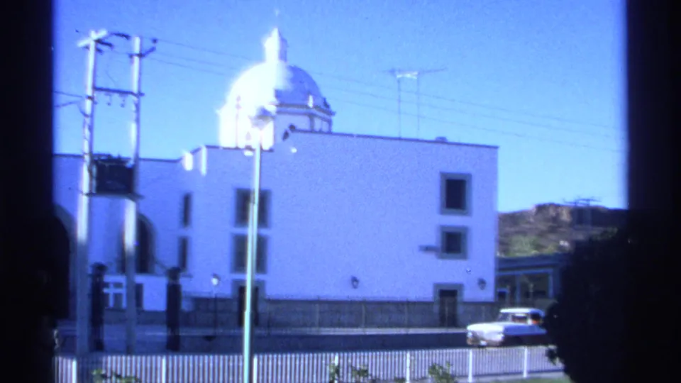 Exterior 1975 Guaymas view of a white domed church beside a street, with railings, utility poles, and a light-colored car nearby.
