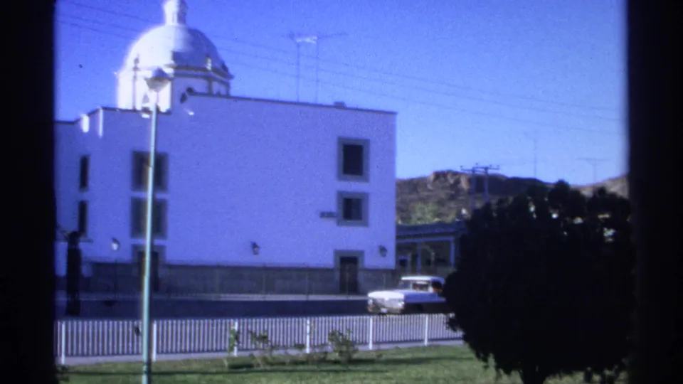 1975 Guaymas exterior frame shows the white church facade and dome, street railings, a car, trees, and low hills in the background.