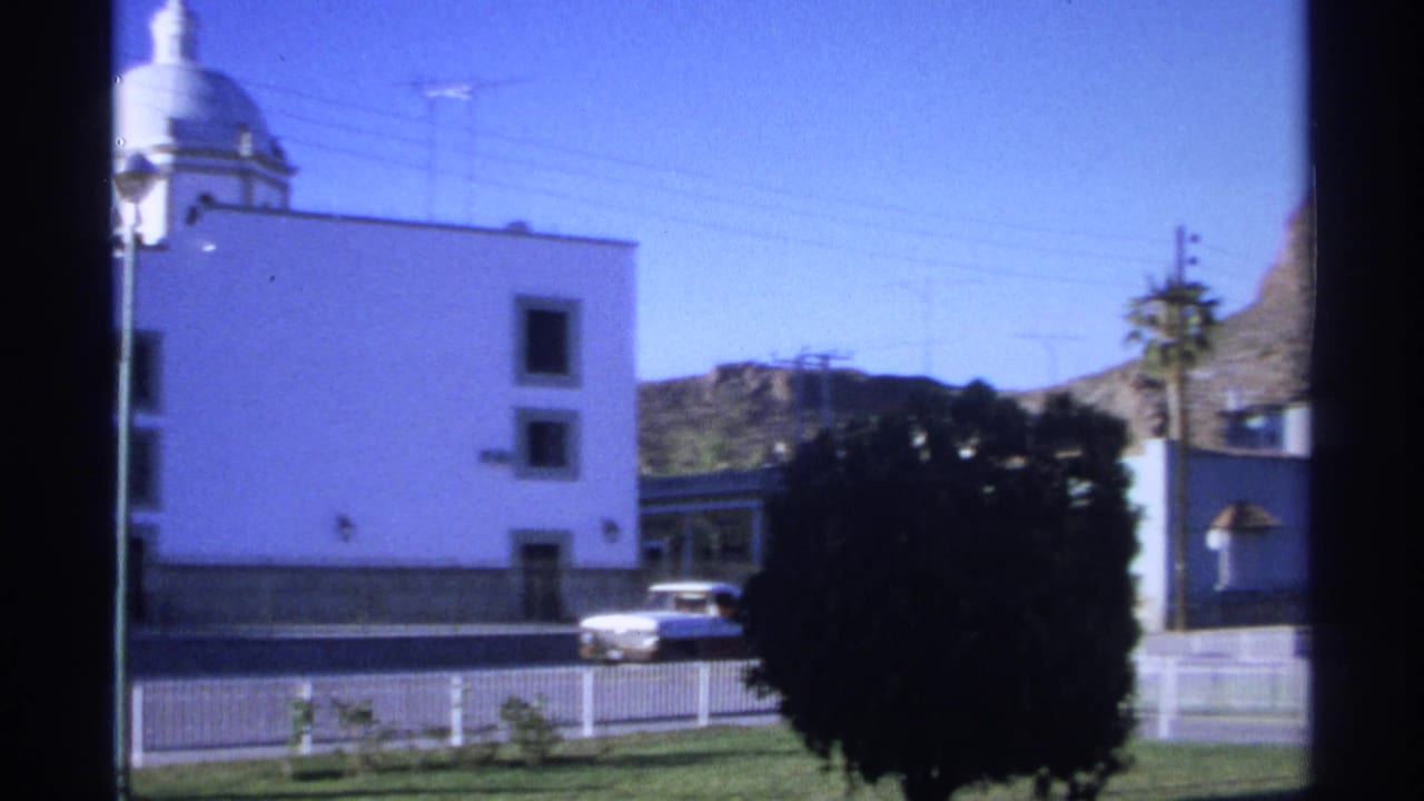 White building with domed roof, dark bush, white pickup, fence, and mountain beyond.
