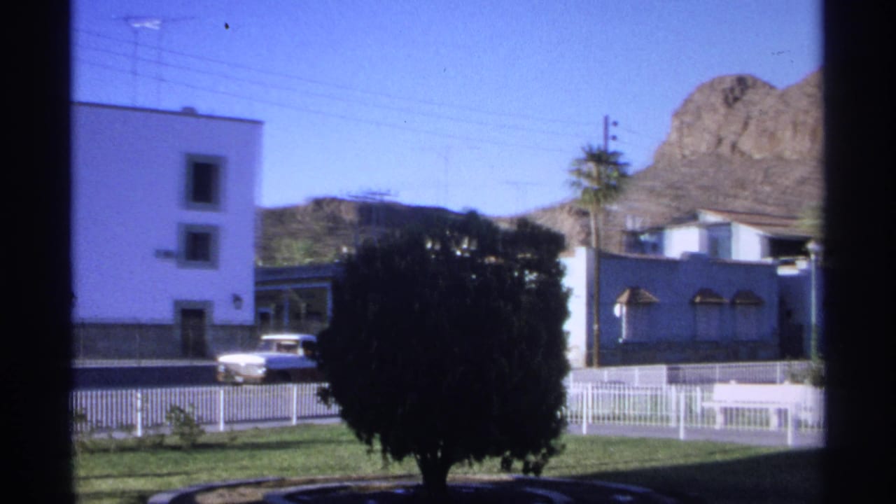 White and blue buildings, white pickup, dark bush, palm tree, fence, and rocky hill.