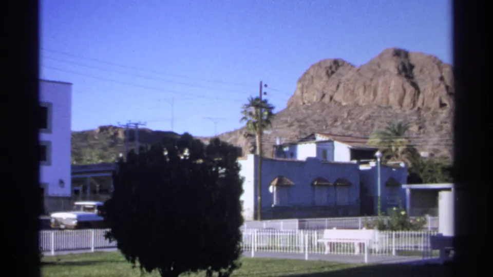 Guaymas 1975 street pan with a rocky mountain rising behind low buildings, a palm tree, dark foreground bush, and white fence.