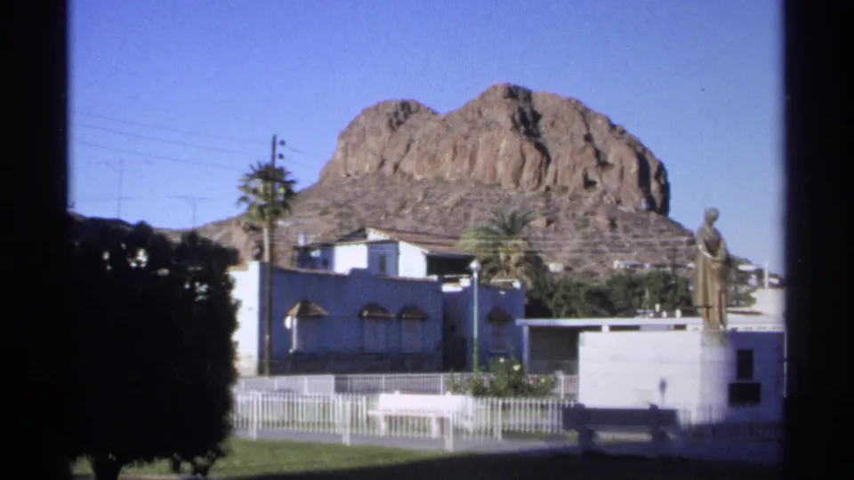 1975 Guaymas street view with rocky mountain backdrop, blue building, palm tree, white fence, dark shrub, and statue on a pedestal.