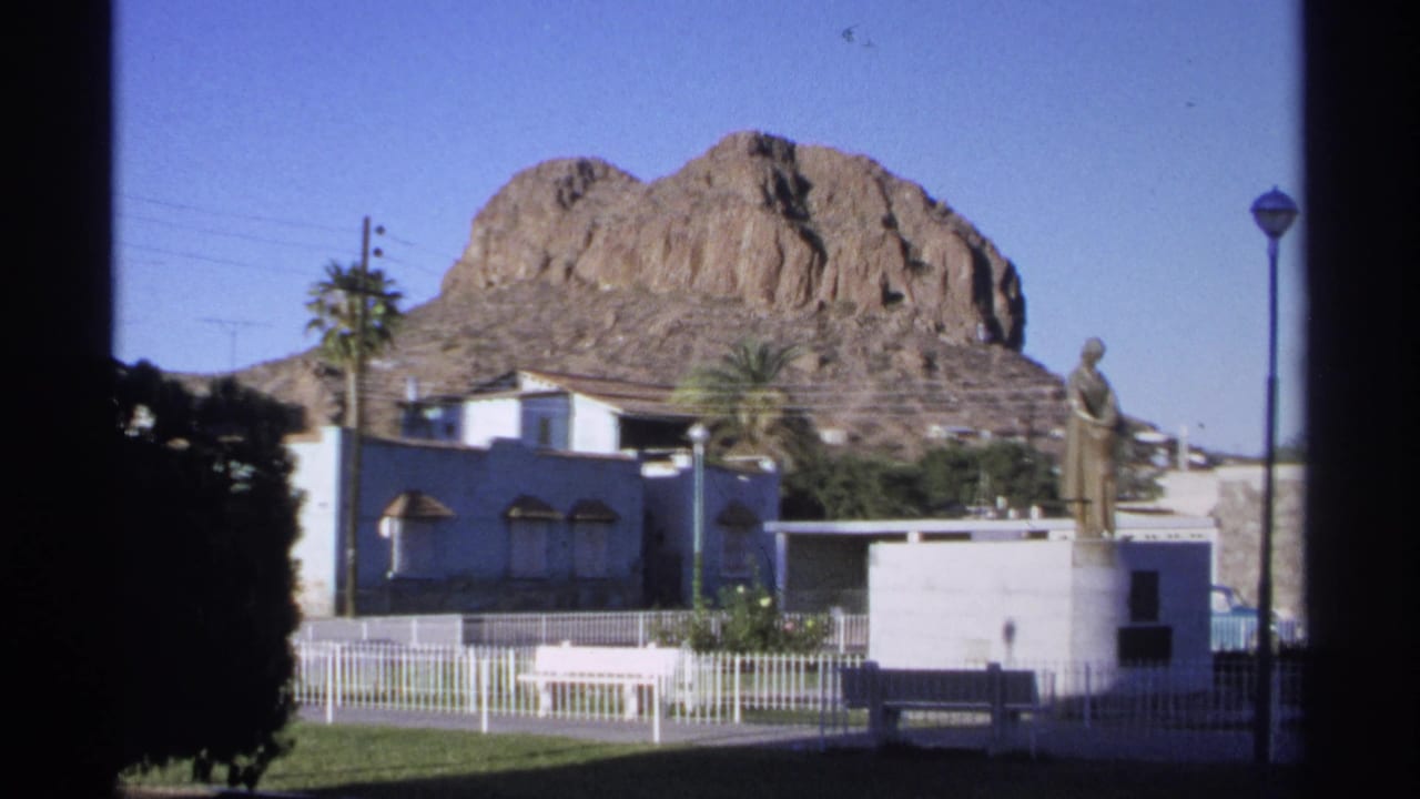 Bronze statue on a white pedestal in a fenced plaza with palm trees and a rocky hill behind it.