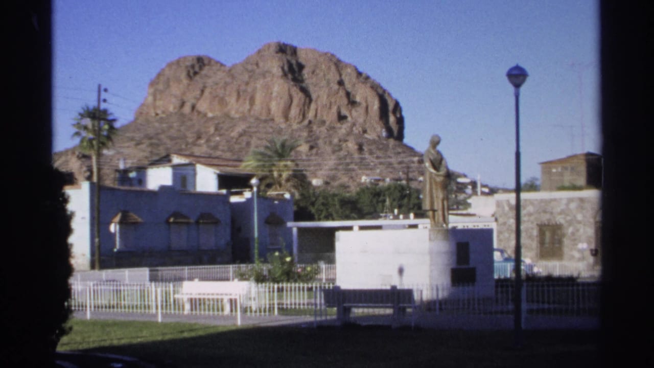 Plaza statue, white fence, benches, palms, and rocky mountain seen under a clear sky.