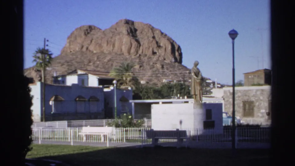Guaymas, Mexico plaza view from 1975 8mm footage, showing a statue on a pedestal with palms, fencing, buildings, and a rocky hill.