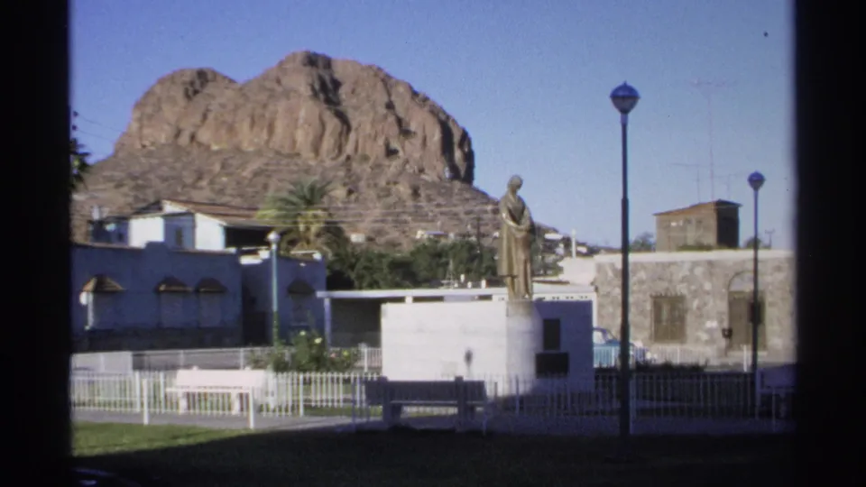 1975 visitor footage in Guaymas shows the plaza monument framed by a tall streetlamp, white fence, bench, buildings, and rocky hillside.